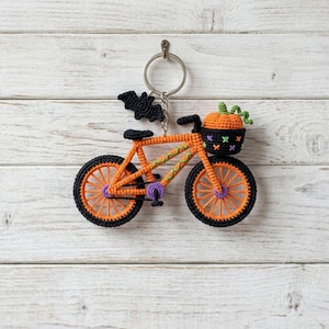 Puede incluir: Un llavero de ganchillo con forma de bicicleta naranja con ruedas negras, un pedal morado y un murciélago negro unido a la parte superior. Una pequeña calabaza naranja con tallo verde descansa en una cesta negra sobre el manillar, decorada con pequeños patrones de punto de cruz morados y verdes.