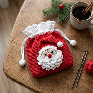 Puede incluir: Una bolsa de ganchillo roja con cordón, borde superior festoneado blanco y una aplicación central de cara de Papá Noel con rostro beige, barba blanca y nariz roja. La bolsa incluye cordones trenzados blancos con pequeños pompones.