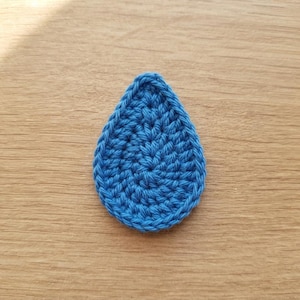 May include: A teardrop-shaped crochet applique crafted from vibrant blue yarn. The piece features a dense, uniform stitch pattern that creates a textured surface, forming a distinct pointed top and a rounded base with a consistent blue color throughout.