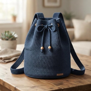 Può includere: Uno zaino a sacco in uncinetto blu scuro con chiusura a coulisse e due perline in legno chiaro alle estremità. La borsa presenta una base cilindrica strutturata, due spallacci e una piccola etichetta rettangolare applicata sul davanti.