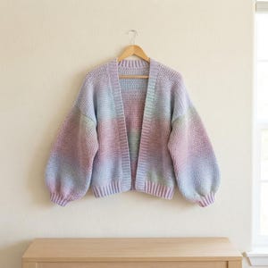 May include: A long-sleeved open-front cardigan made of crocheted yarn featuring a soft gradient of pastel colors including light blue, lavender, pale pink, and mint green. The garment has a ribbed collar, cuffs, and hem with voluminous sleeves.