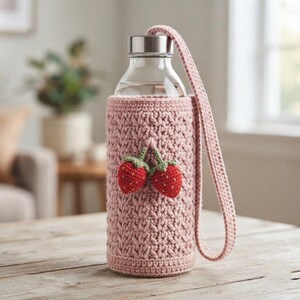Può includere: Una bottiglia d'acqua in vetro trasparente inserita in una custodia all'uncinetto rosa polvere con un motivo a punti testurizzati e due piccole fragole rosse all'uncinetto con steli verdi, dotata di una tracolla coordinata all'uncinetto.