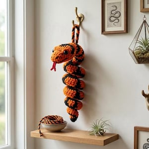 Op de afbeelding: Een gehaakt slangenfiguur met afwisselende oranje en zwarte garensegmenten, een rode tong van garen en donkere plastic ogen. De slang hangt aan een goudkleurige metalen wandhaak, met een opgerold stuk garen in een klein schaaltje op een houten plank.