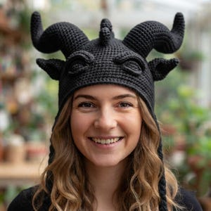 May include: A black crocheted hat featuring two large, curved horns, two smaller ears, and a central ridge detail. The hat is constructed with a dense, textured stitch pattern and includes long, braided tassels hanging down from the sides.