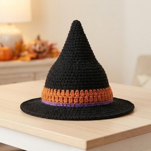 May include: A pointed witch hat crafted from black yarn with a textured crochet stitch. The base of the cone features a wide band of orange yarn followed by a thin stripe of purple yarn, encircling the crown above the flat, circular brim.