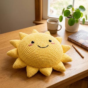 Könnte beinhalten: Ein hellgelbes, gehäkeltes Sonnenkissen mit rundem Mittelteil und dreieckigen Strahlen. Das Gesicht hat zwei schwarze Kunststoffaugen, einen aufgestickten schwarzen geschwungenen Mund und zwei kleine rosa Wangen auf texturierter Wolle.