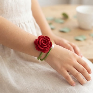 May include: A wrist accessory featuring a deep red crocheted rose flower attached to a green wrapped band. The band includes a single white spherical bead accent. The design is constructed from yarn and thread, showcasing a floral motif on a flexible cord.