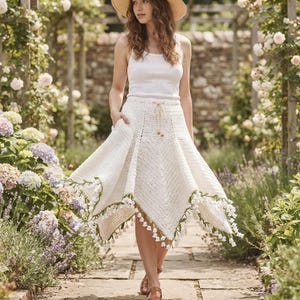 May include: A white sleeveless tank top paired with a cream-colored crochet skirt featuring a drawstring waist and a handkerchief hemline adorned with green vine-like accents and small white bell-shaped floral embellishments along the edges.