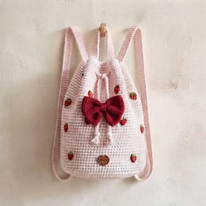 Può includere: Uno zaino con coulisse realizzato all'uncinetto in rosa chiaro, caratterizzato da un grande fiocco rosso scuro al centro e piccoli motivi a fragola ricamati sulla superficie. La borsa include due spallacci e un'etichetta metallica rotonda.