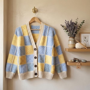 Puede incluir: Un cárdigan de ganchillo con un patrón de cuadros en tonos azul claro y amarillo pálido. La prenda presenta un cuello en V, mangas largas, puños y dobladillo acanalados en color crema, y cuatro botones de color marrón oscuro en el frente.