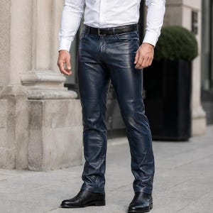 Puede incluir: Una camisa blanca de manga larga con cuello clásico, combinada con pantalones de cuero azul marino oscuro que presentan una costura horizontal sobre la rodilla, un cinturón de cuero negro y botas de vestir de cuero negro.