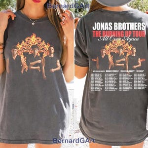 May include: A charcoal grey short-sleeved t-shirt featuring a graphic of the Jonas Brothers in front of a flame design. The back displays the text Jonas Brothers The Burning Up Tour All Over Again with a list of tour dates and locations.