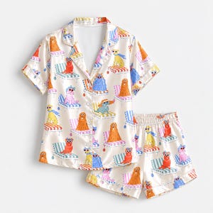 May include: A cream-colored pajama set consisting of a short-sleeved button-down shirt and matching shorts. The fabric features a repeating pattern of various dogs in sunglasses and hats lounging on striped beach chairs in shades of red, blue, and pink.