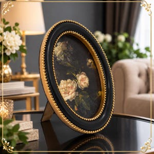 May include: An oval picture frame with a black outer border and a gold beaded inner trim. The frame contains a dark print featuring light-colored roses and green leaves. It stands on a support leg, displaying a classic design with high contrast.