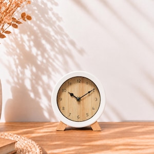 May include: A round analog table clock featuring a white frame and a light wood grain face. The clock has black numerals from 1 to 12 and two black hands. It is supported by two small, light-colored wooden feet at the base.