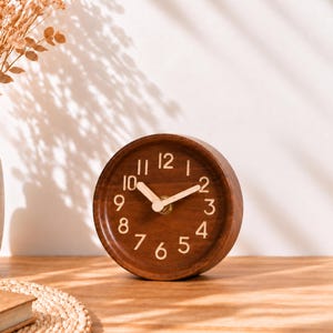 May include: A round wooden analog clock with a dark brown finish, featuring light-colored numerals from 1 to 12 and matching light-colored hour and minute hands. The clock face is simple and circular, showcasing a natural wood grain texture.