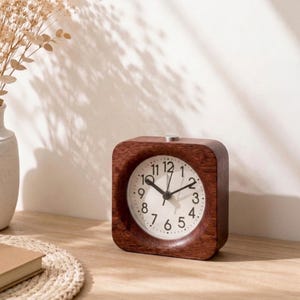 May include: A square alarm clock with a dark brown wood frame and a white circular clock face. The face features black Arabic numerals from 1 to 12, black hour, minute, and second hands, and a small silver button on the top of the wooden casing.