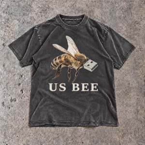 May include: A charcoal gray short-sleeve t-shirt featuring a graphic of a honeybee with a silver USB flash drive for a head. The text US BEE is printed in a cream-colored serif font below the bee illustration on the front of the garment.