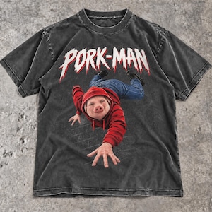 Puede incluir: Una camiseta gris carbón de manga corta con el gráfico de una figura con cara de cerdo que viste una sudadera roja con capucha y vaqueros azules. La figura extiende una mano. El texto PORK-MAN está impreso en blanco sobre la figura.