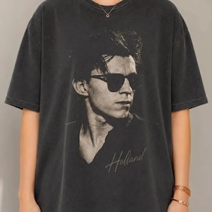 Może przedstawiać: Ciemnoszary t-shirt z krótkim rękawem z czarno-białym nadrukiem graficznym przedstawiającym osobę w okularach przeciwsłonecznych. Nadruk zawiera napis Holland wykonany kursywą pod portretem. Koszulka posiada okrągły dekolt pod szyją.