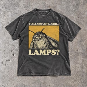 Może przedstawiać: Ciemnoszary t-shirt z krótkim rękawem z grafiką ćmy umieszczoną na żółtym prostokątnym tle. Tekst nad ćmą brzmi Y'ALL GOT ANY.. UHH..., a tekst pod ćmą brzmi LAMPS? i jest napisany jasną czcionką o pogrubionym kroju pisma.