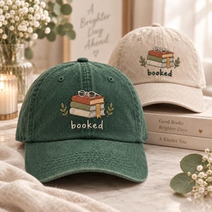 Könnte beinhalten: Eine dunkelgrüne Baseballkappe und eine beige Baseballkappe, beide mit einer Stickerei eines Bücherstapels aus drei Büchern, einer Brille darauf und dem Wort booked darunter, flankiert von zwei kleinen grünen Blattzweigen auf jeder Seite.