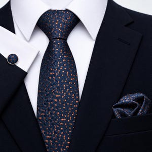 Peut inclure: Une cravate bleu marine foncé avec un motif texturé à taches cuivrées, accompagnée d'une pochette assortie et d'un bouton de manchette circulaire, le tout porté avec une chemise blanche et une veste de costume bleu marine uni.