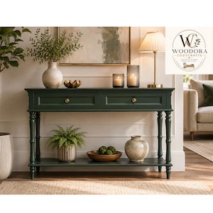 May include: A dark green wooden console table with two drawers, brass knobs, and a lower shelf. The table features turned legs and a rectangular top. The Woodora Cozycrafts logo is visible in the upper right corner of the image.