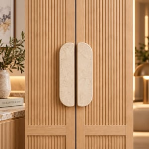 Peut inclure: Une paire de portes de placard en bois clair à lattes verticales, dotées de deux poignées allongées en demi-cercle en pierre beige tachetée. Les portes présentent une texture nervurée uniforme sur toute leur surface.