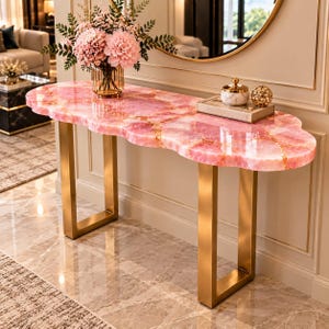May include: A console table featuring a polished pink quartz slab top with irregular, organic edges and gold-toned metal legs. A glass vase with pink flowers, a small lidded jar, and a decorative gold sphere sit on the surface.