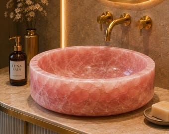 Puede incluir: Un lavabo de sobreponer redondo de cuarzo rosa pulido con una textura cristalina natural. El cuenco se asienta sobre una encimera de mármol junto a un dispensador de jabón de vidrio marrón con la etiqueta LUNA SAIN y una jabonera.