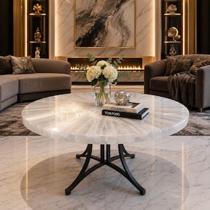May include: A round coffee table with a white, sunburst-patterned stone top and a black metal base. On the table sits a clear glass vase with white hydrangea flowers, a silver decorative sphere, and two stacked books labeled Chanel and Tom Ford.