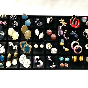 Puede incluir: Una bandeja expositora de joyas rectangular de color negro que contiene una colección de pendientes variados de diversas formas, colores y diseños, incluyendo broqueles, aros y colgantes, organizados en compartimentos cuadrados individuales.