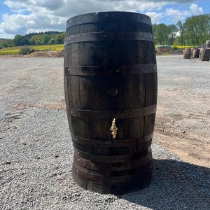 May include: A dark brown wooden barrel featuring multiple horizontal metal bands and a brass spigot installed on the lower front section. The cylindrical container has a weathered texture and is constructed from vertical wooden staves.
