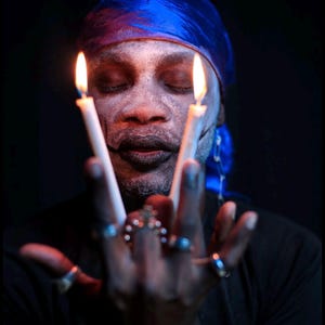 May include: A person wearing a bright blue durag and dark makeup with white face paint, holding two lit white taper candles. The person has multiple silver rings on their fingers and a silver safety pin earring, set against a dark background.