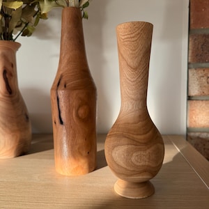 May include: Three turned wooden vases with distinct grain patterns and smooth finishes. The front vase features a rounded base and a slender neck, while the two taller vases behind it have cylindrical shapes and contain dried foliage.