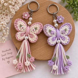 Puede incluir: Dos llaveros de mariposa tejidos a ganchillo con anillas metálicas. Uno es rosa y crema, el otro es morado y crema. Ambos tienen una cuenta de perla central, un pequeño dije de flor tejida y una borla de hilo larga con flores diminutas.