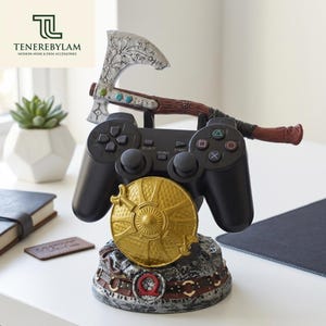 May include: A black video game controller stand featuring a silver-colored axe with intricate engravings, a gold-toned circular shield, and a textured base with a red circular emblem. The stand is designed to hold a controller securely in place.