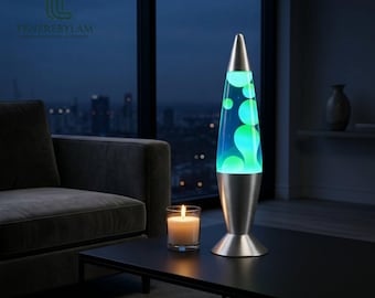 May include: A cone-shaped lava lamp with a brushed silver metal base and cap, containing a blue liquid with glowing green wax blobs. The lamp is positioned next to a small, lit white pillar candle in a clear glass holder on a black table.