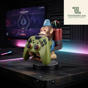 May include: A figurine of a monkey wearing a blue fez hat and carrying red dynamite sticks on its back, holding an olive green video game controller. The controller features black joysticks, a directional pad, and four colored action buttons.