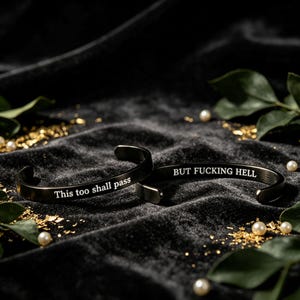 May include: Two black metal cuff bracelets with engraved white text. One bracelet features the phrase "This too shall pass" and the other features the phrase "BUT FUCKING HELL". The bracelets have a smooth, matte finish and an open-ended design.
