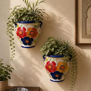 May include: Two white ceramic wall planters featuring hand-painted floral motifs in red, orange, and blue. Each vessel has two side handles and contains green foliage. The planters are mounted on a wall, with a small matching bowl on a table.