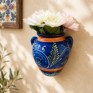 May include: A wall-mounted ceramic vase featuring a deep blue glaze with hand-painted yellow, orange, and white floral scrollwork patterns. The vessel has two side handles, an orange rim, and contains a bouquet of white and light pink dahlia flowers.