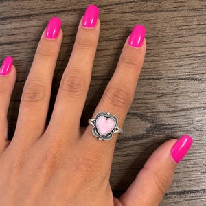 May include: A silver ring featuring a central heart-shaped light pink stone. The metal setting has a decorative scalloped edge with small bead accents surrounding the stone. The band is smooth and polished, showcasing a detailed metalwork design.