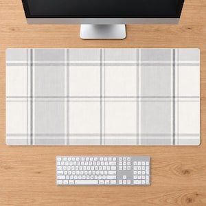 May include: A rectangular desk mat featuring a light gray and white plaid pattern with intersecting vertical and horizontal lines. The mat is positioned on a wooden surface below a computer monitor and above a white computer keyboard.