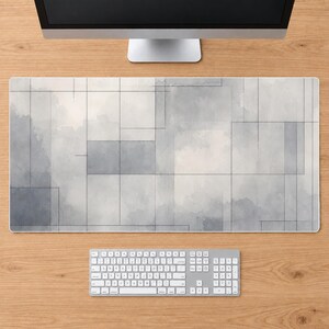 Puede incluir: Una alfombrilla de escritorio rectangular con un patrón abstracto inspirado en la acuarela en tonos gris, blanco y carbón. El diseño consiste en formas rectangulares superpuestas y líneas finas oscuras que crean una cuadrícula geométrica.