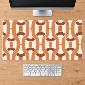 Peut inclure: Un tapis de bureau rectangulaire présentant un motif géométrique répétitif de formes enchaînées entrelacées dans des tons orange brûlé, terre cuite et crème. Le tapis est placé sur une surface en bois sous un écran et un clavier blanc.