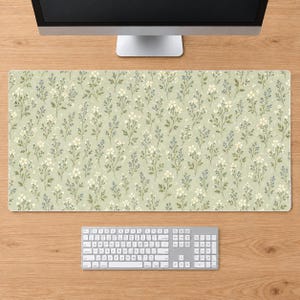 Puede incluir: Una alfombrilla de escritorio rectangular con un patrón repetitivo de pequeñas flores blancas, hojas verdes y diminutas ramitas azules sobre un fondo verde salvia claro. La alfombrilla está colocada sobre una superficie de madera bajo un monitor.