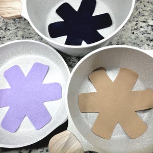 May include: Three white cooking pots with wooden handles, each containing a felt star-shaped insert in navy blue, lavender, and tan. The pots are arranged on a speckled countertop, showcasing a kitchenware set.