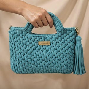 May include: A teal crochet handbag featuring a thick, textured knit pattern with two integrated handles. A rectangular gold-toned metal tag with the word Handmade is centered on the front, and a matching teal fabric tassel hangs from the side.